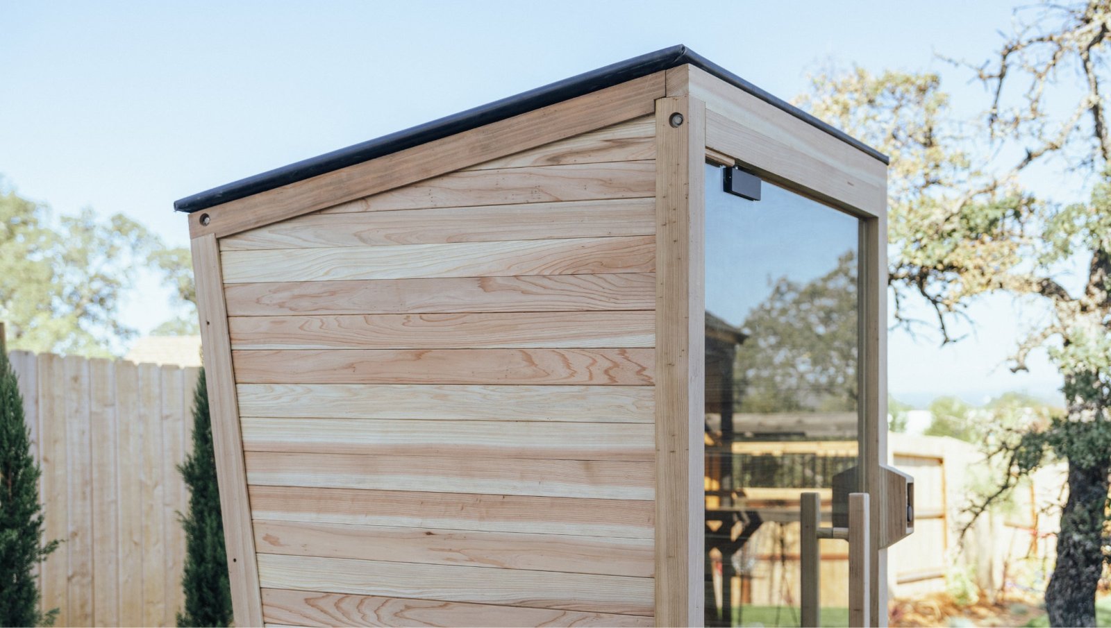 The Plunge Sauna Mini, featuring HUUM heater and glass door, stands in a fenced backyard with an angled roof.