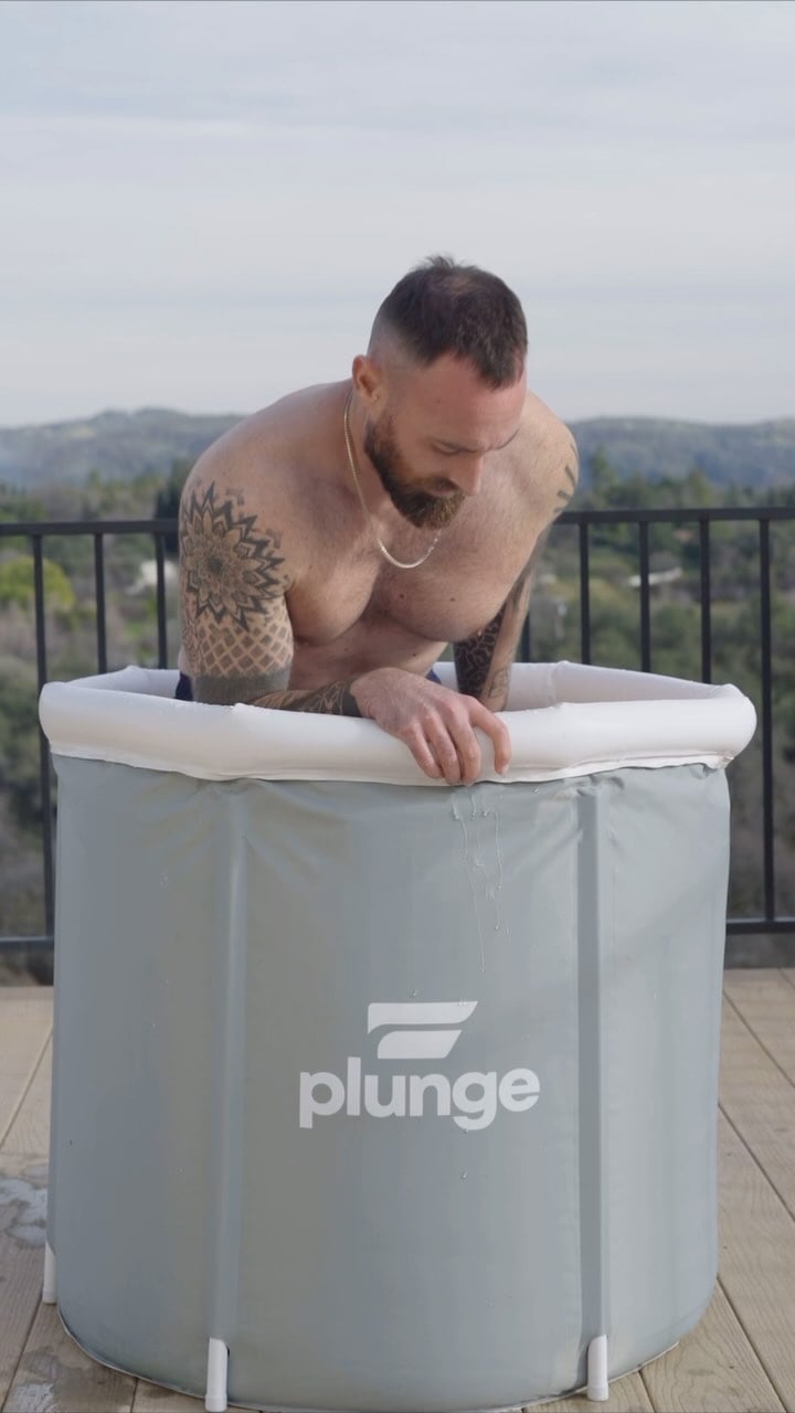 Man with tattoos entering an outdoor ice bath labeled 'plunge.'