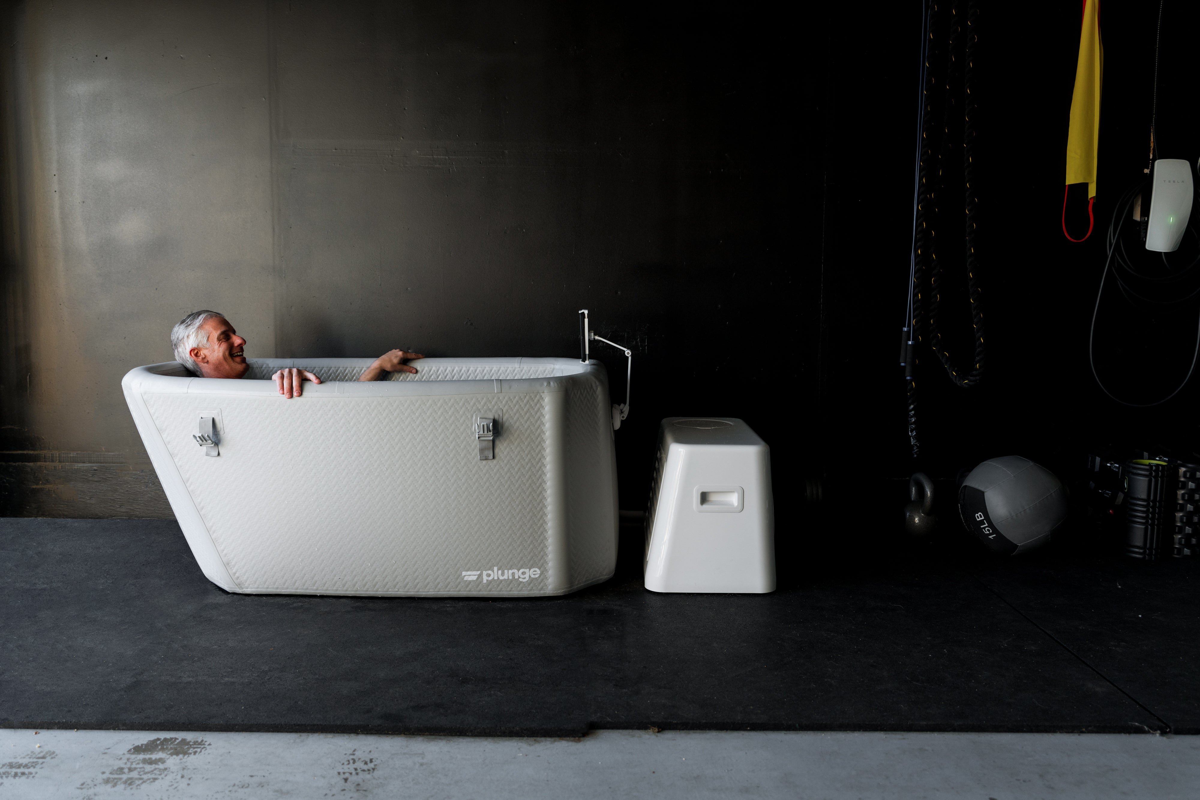 A person enjoys an ice bath in a white plunge tub inside a dark gym-like setting.