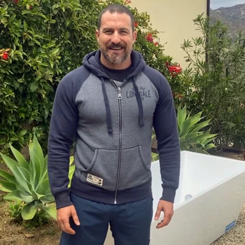 Man in a grey hoodie smiling in a garden with greenery and plants.