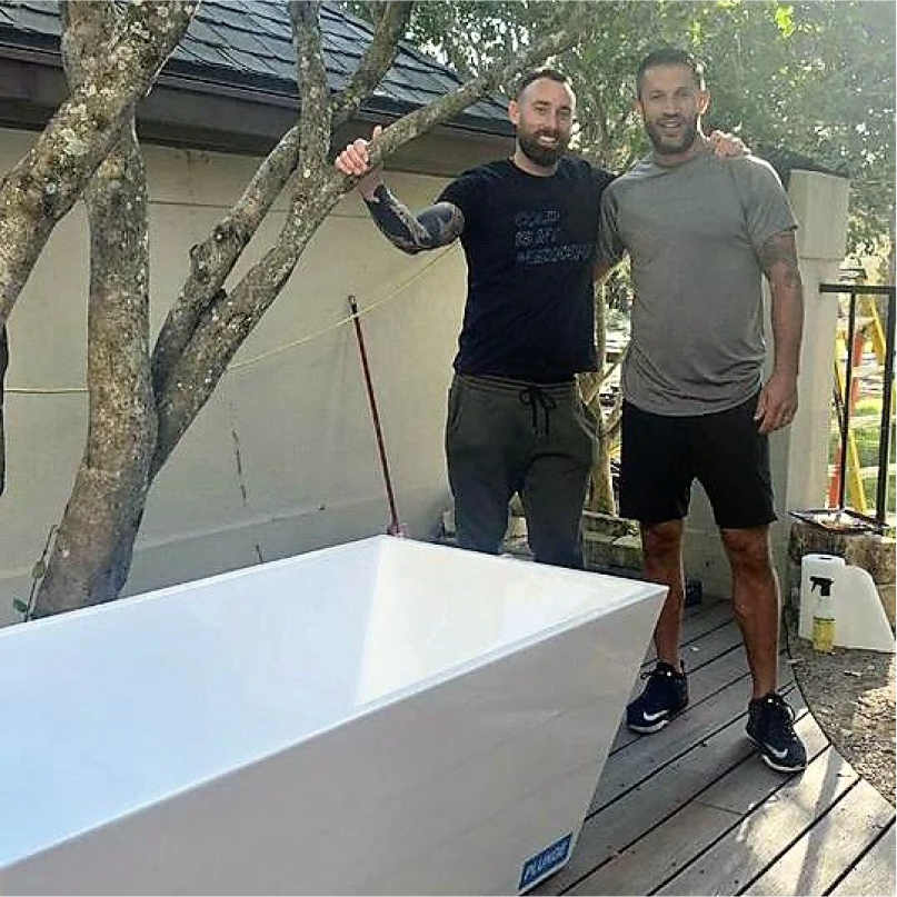 Two men standing next to a white bathtub on a wooden deck outdoors.