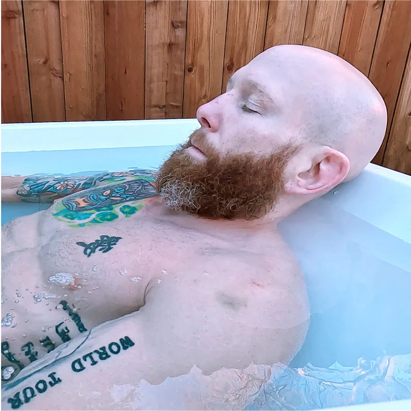 Bearded man with tattoos relaxing in an outdoor bathtub.