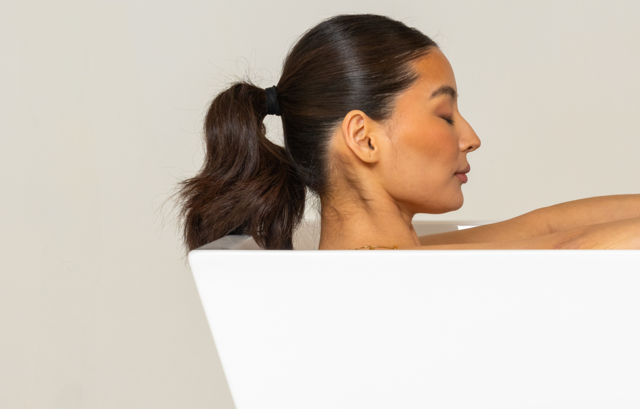 Profile of a woman with closed eyes, in a white bathtub, hair tied in a ponytail.