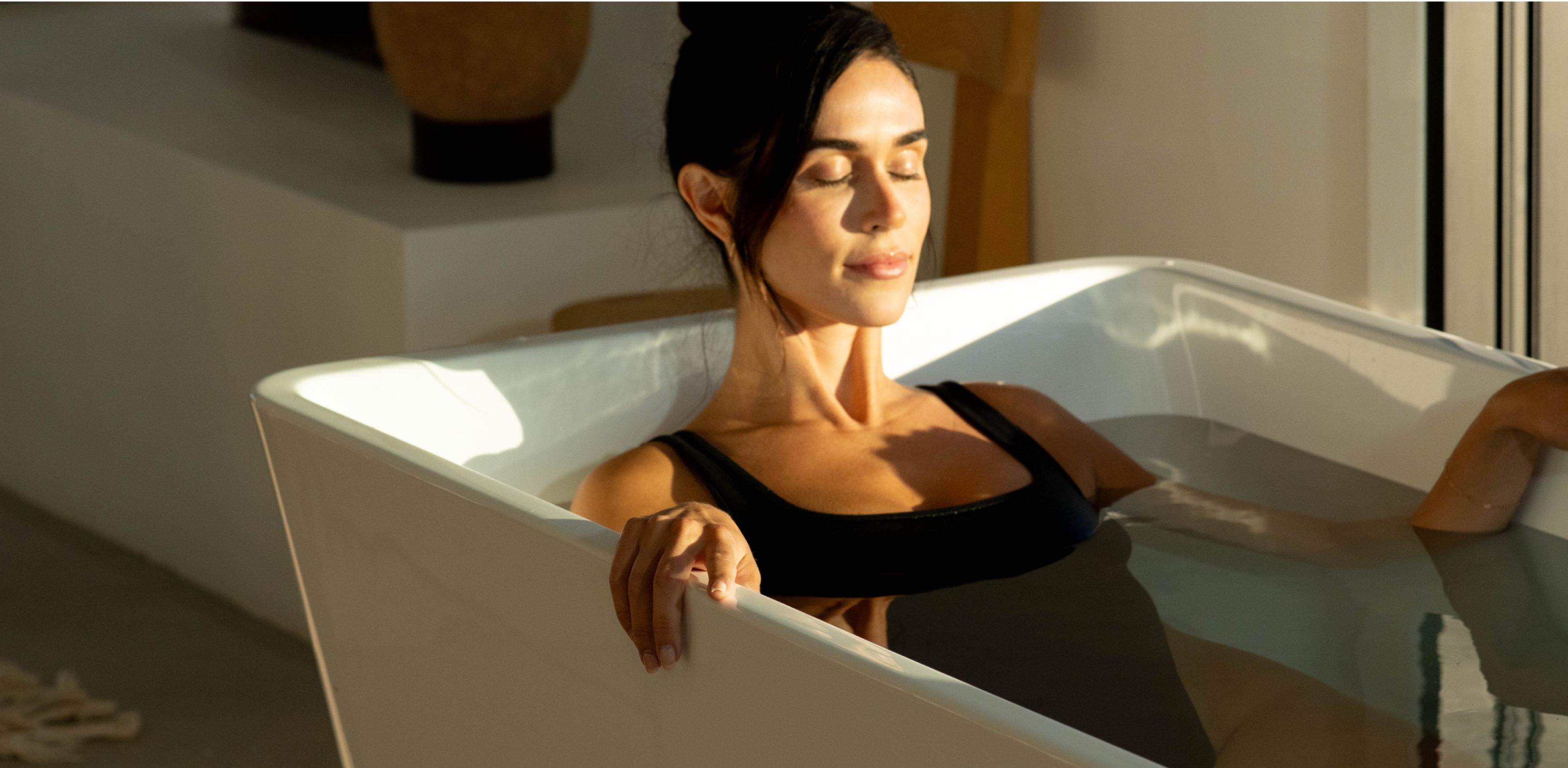 A man with a beard and gold chain stands in a bathtub with his eyes closed.