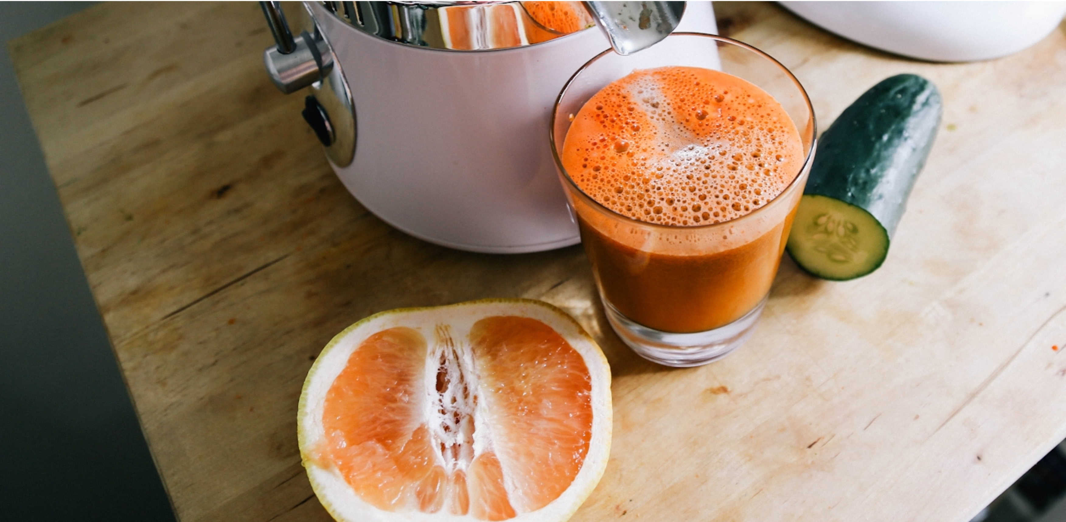 A fresh glass of juice placed on a wooden table beside halved citrus fruit and a cucumber.