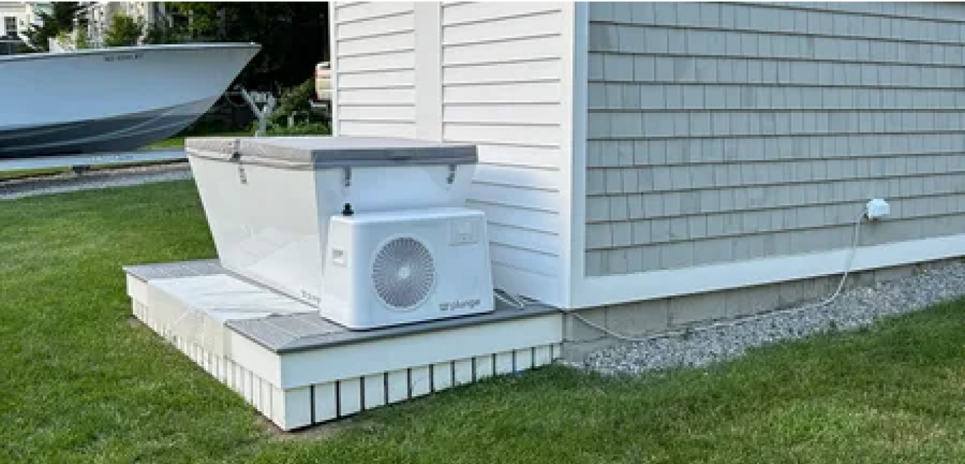 Air conditioner unit installed on a small platform outside a house with a boat in the background.