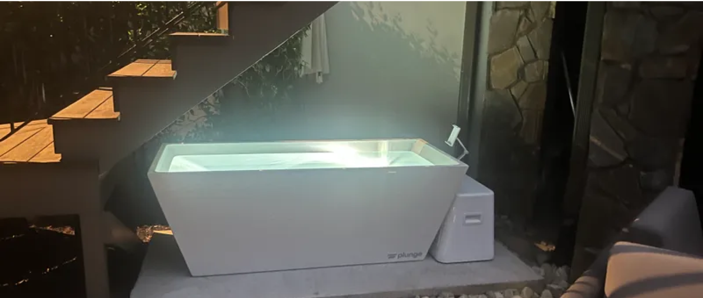 Outdoor bathtub setup under wooden stairs, next to a stone wall.